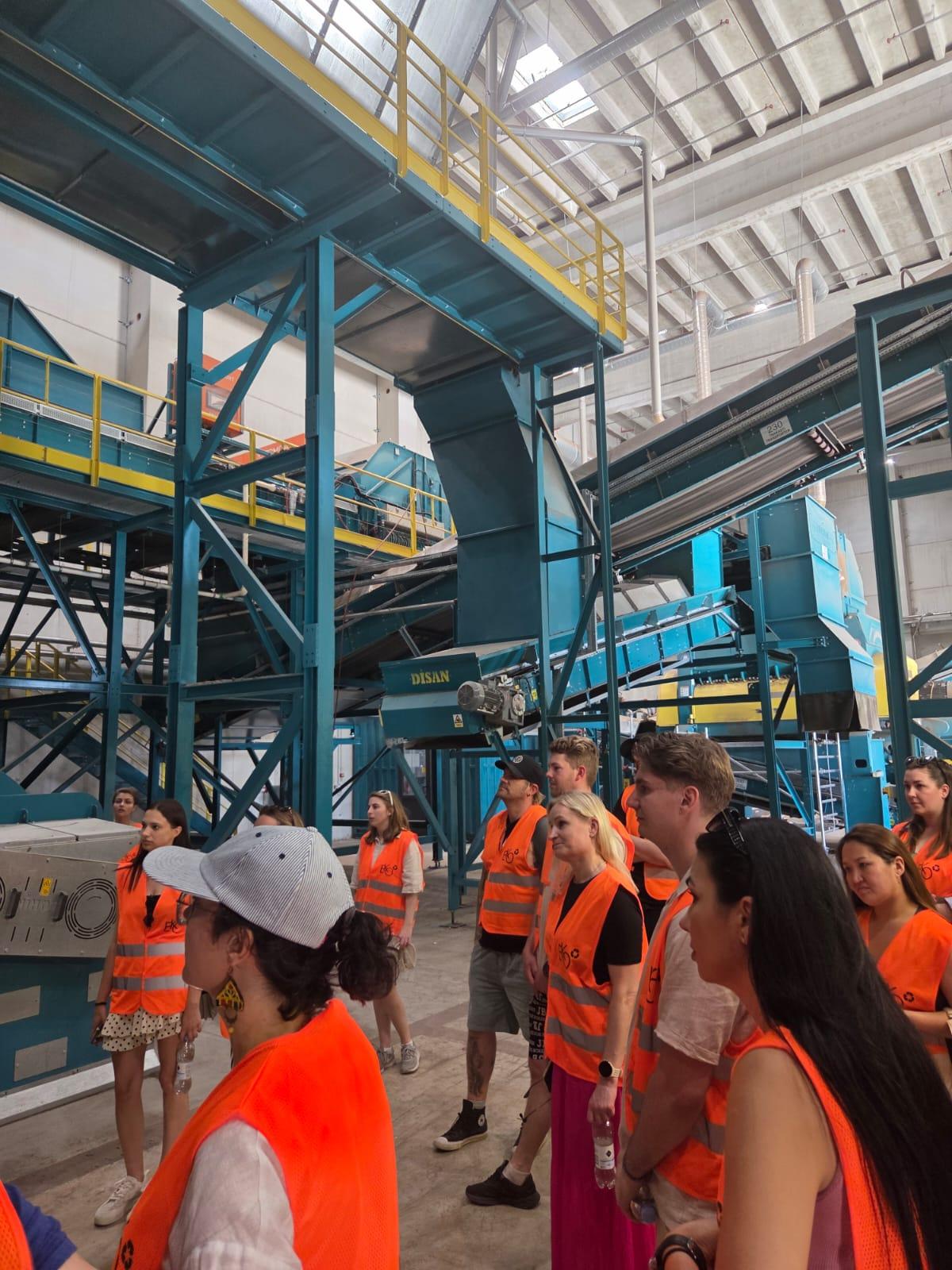 Eine Gruppe von Personen mit orangefarbenen Sicherheitswesten und Mützen besichtigt eine industrielle Recyclinganlage mit großen blauen Maschinen, Förderbändern und hohen Metallstegen unter einer hellen, hohen Decke.