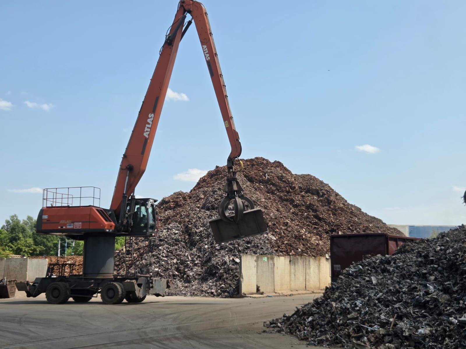 Ein großer orangefarbener Bagger mit einem Krallenaufsatz hebt an einem sonnigen Tag Schrott vor einem großen Haufen Metallschrott auf einem Recycling- oder Schrottplatz auf.