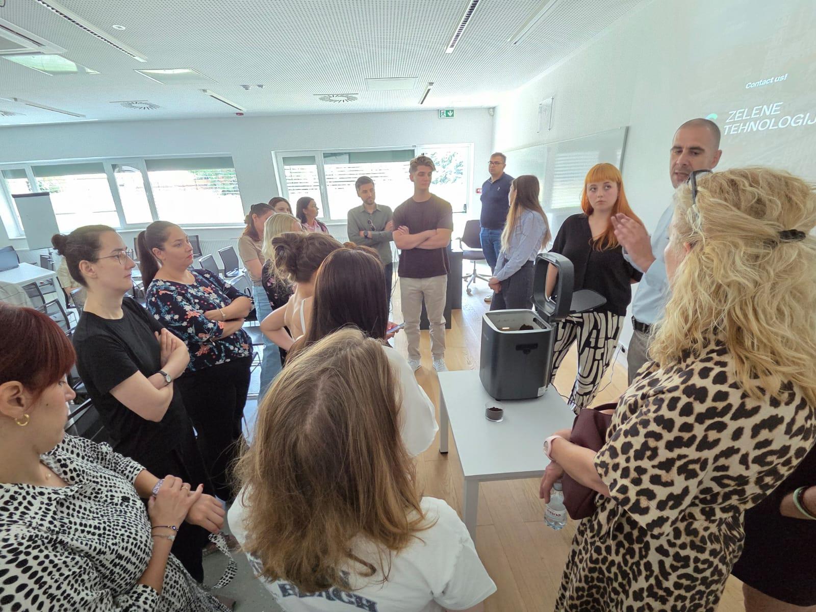 Eine Gruppe von Menschen steht in einem hellen Klassenzimmer, versammelt um einen Tisch, an dem ein Mann etwas über ein Gerät erklärt. Alle schauen aufmerksam zu, und an der Wand hinter ihnen wird eine Präsentation projiziert.