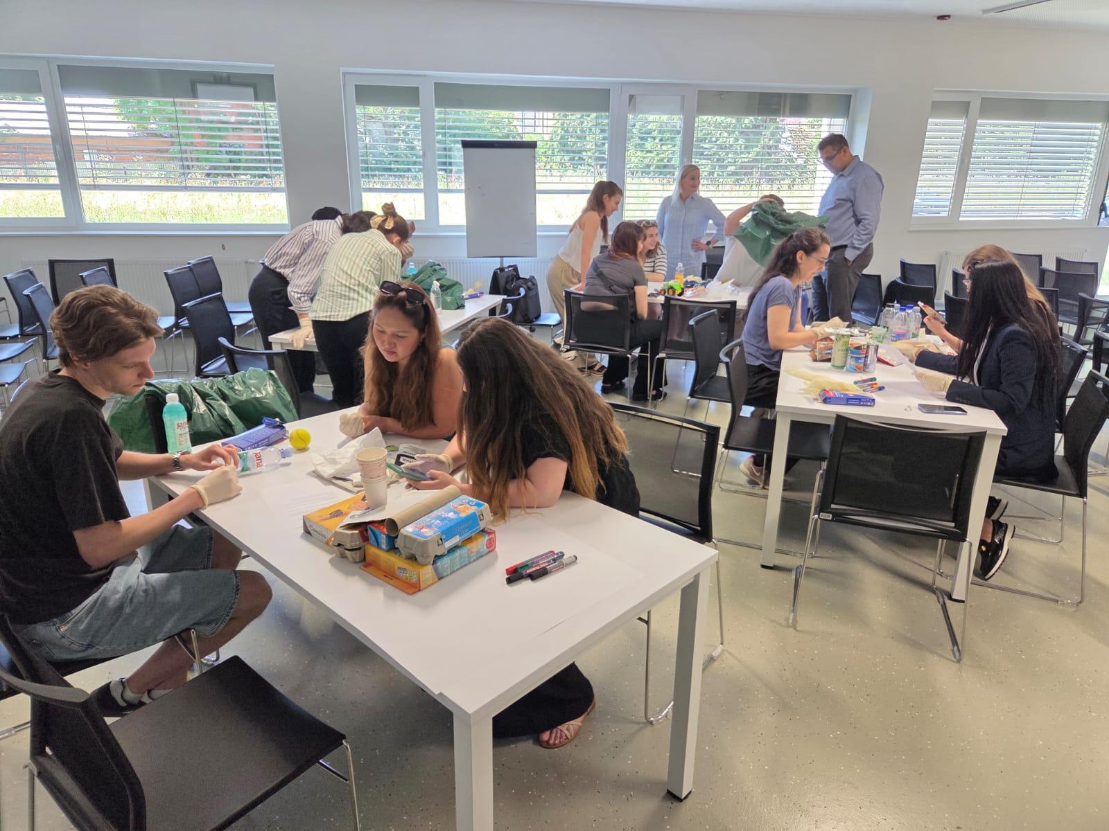 Eine Gruppe von Personen sitzt an Tischen in einem hellen Raum und arbeitet an einem Projekt, bei dem sie verschiedene Lebensmittelbehälter, Dosen und Bastelmaterialien verwenden. Einige Personen stehen im Hintergrund und ordnen das Material.