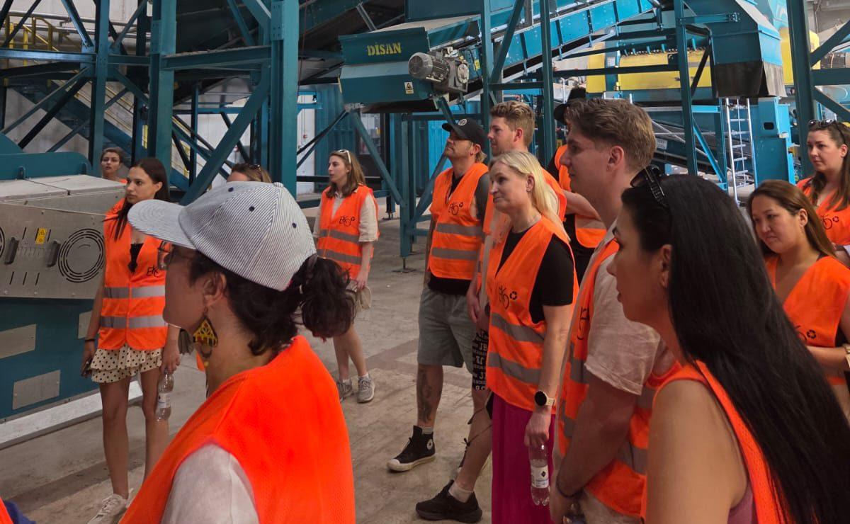 Eine Gruppe von Personen in orangefarbenen Sicherheitswesten und Freizeitkleidung hört bei einer Führung durch eine Industrieanlage mit blauen Maschinen im Hintergrund aufmerksam zu.