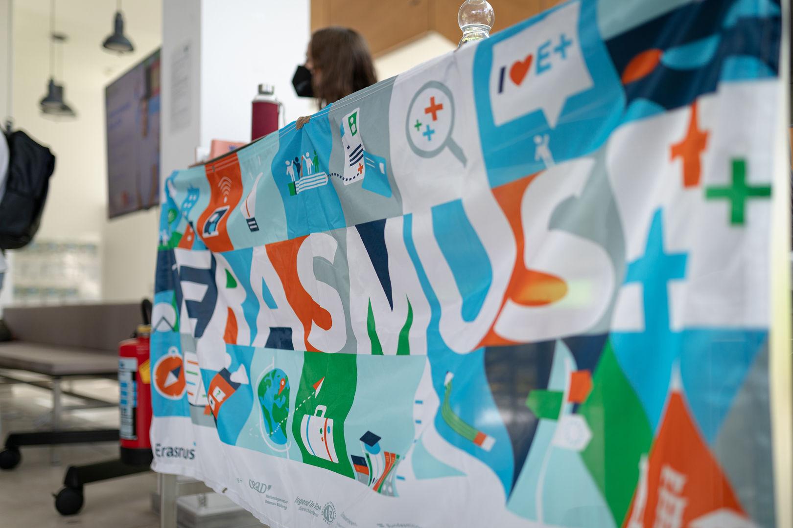 Buntes Erasmus-Banner mit verschiedenen Icons und Symbolen ist über einem Tisch in einem Innenraum drapiert. Auf dem Tisch stehen Wasserflaschen und andere Gegenstände, im Hintergrund sind unscharfe Menschen und moderne Möbel zu sehen.