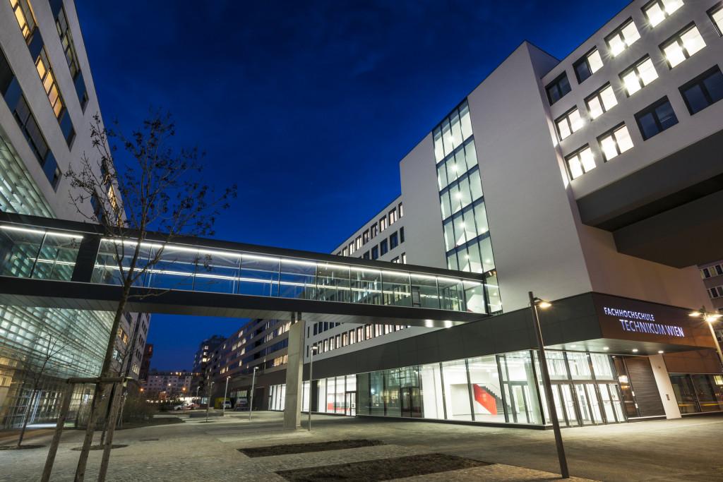 Moderne Hochschulgebäude mit großen Fenstern und einer gläsernen Skybridge, die zwei Gebäude miteinander verbindet, beleuchtet in der Abenddämmerung. Auf dem Schild steht FH Technikum Wien, und der Platz davor ist meist leer.