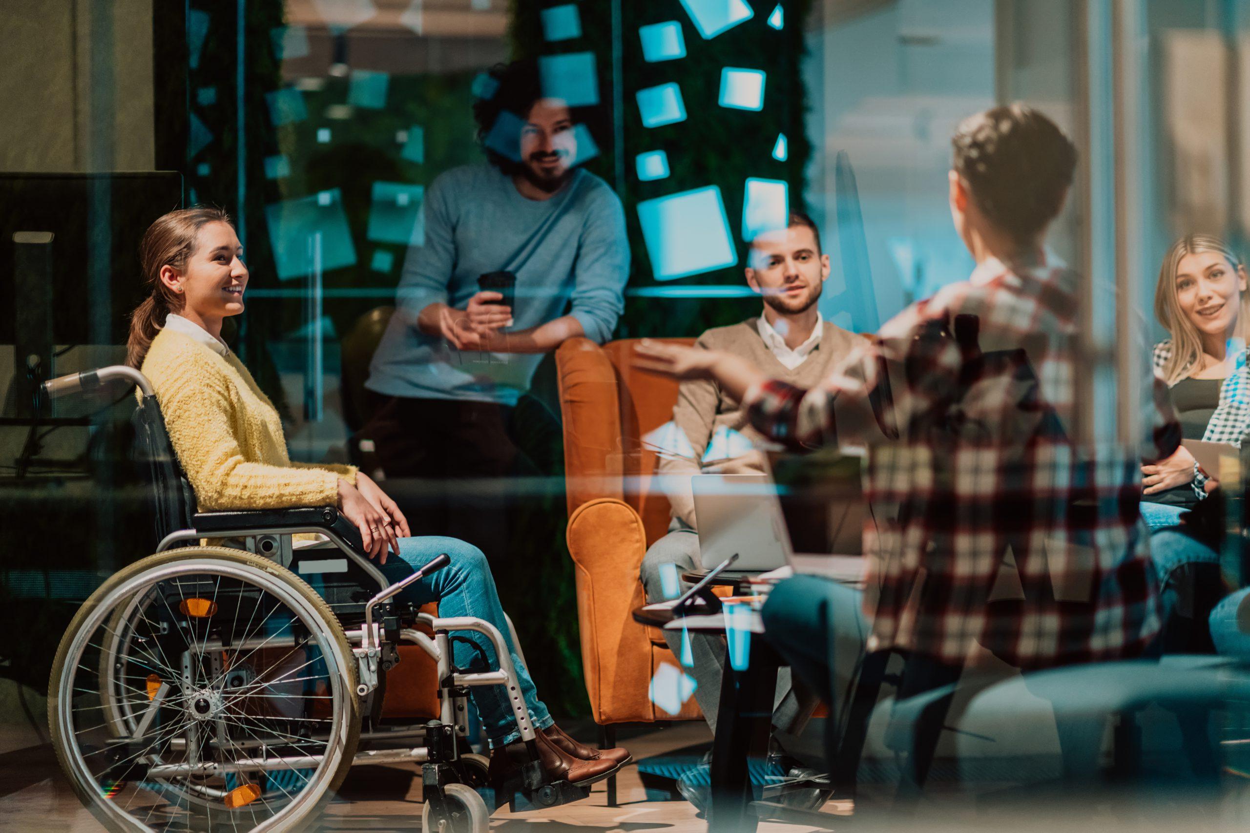 Eine Gruppe von fünf Personen, darunter eine Frau im Rollstuhl, sitzt in einem modernen Büro mit Glaswänden zusammen und führt eine lebhafte Diskussion. Alle wirken engagiert und lächeln.