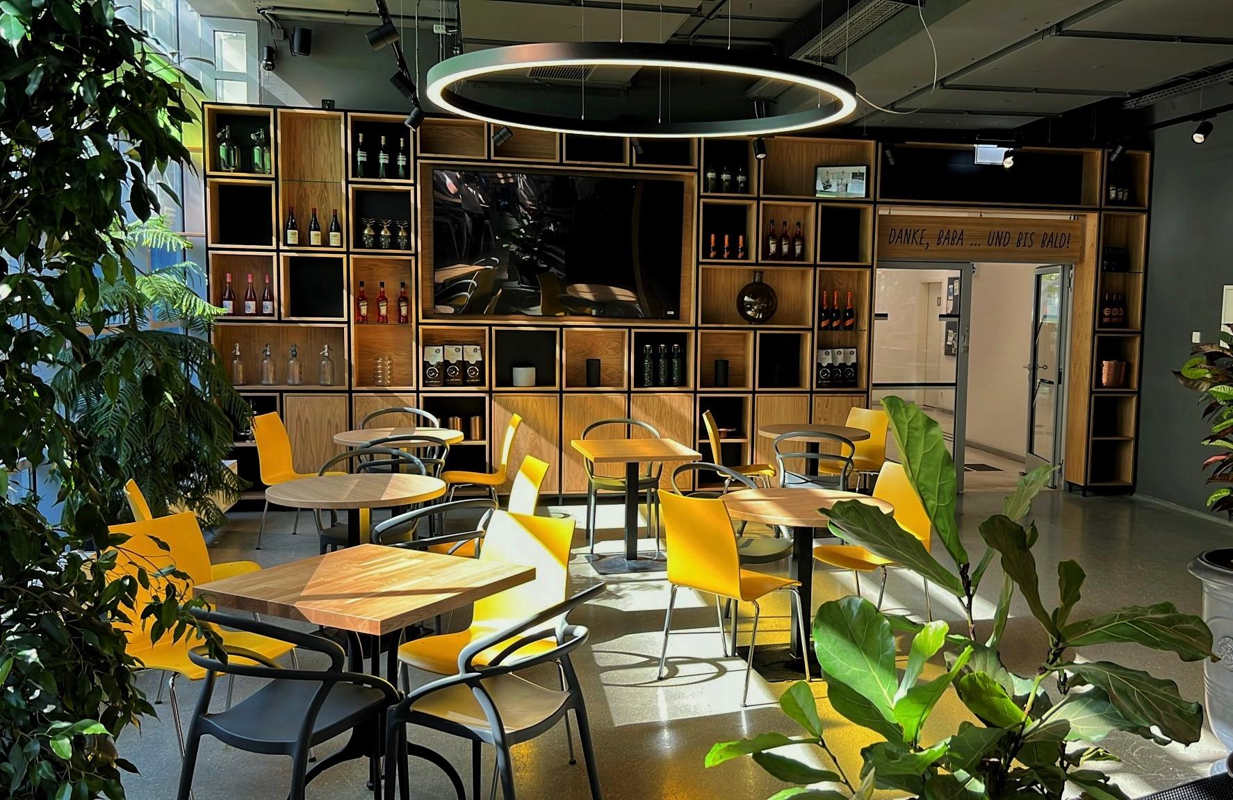 Ein modernes Café mit Holztischen, gelben Stühlen, Grünpflanzen und einer Wand mit Regalen, in denen Flaschen und Dekoration ausgestellt sind. Eine große runde Leuchte hängt von der Decke, und durch die Fenster strömt Sonnenlicht.