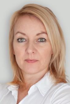 Eine Frau mit hellblondem Haar und grünen Augen blickt direkt in die Kamera. Sie trägt ein weißes Hemd mit Kragen und hat einen neutralen Gesichtsausdruck. Der Hintergrund ist schlicht und hellgrau.