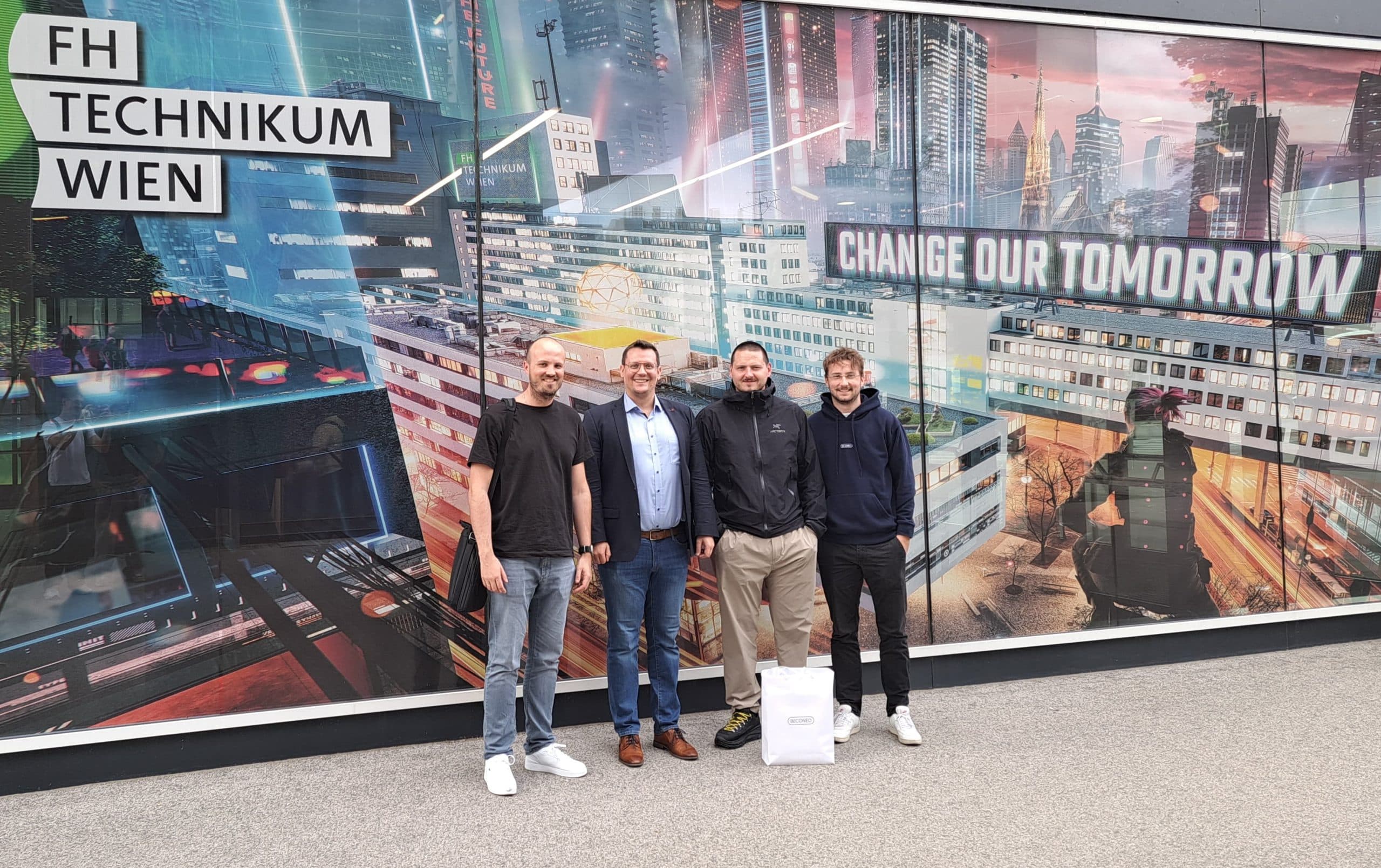 Vier Männer stehen lächelnd vor einem farbenfrohen Wandbild an der FH Technikum Wien, das futuristische Stadtansichten und die Worte 'CHANGE OUR TOMORROW' zeigt. Ein Mann hält eine weiße Tasche in der Hand.