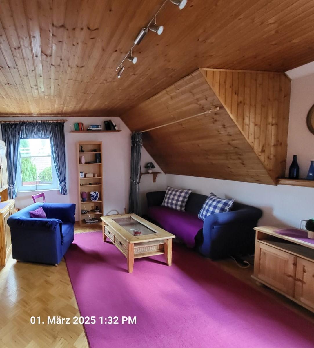 Ein gemütliches Wohnzimmer mit schräger Holzdecke, blauen Sesseln, einem Sofa mit karierten Kissen und lila Bezug, einem hölzernen Couchtisch, Regalen und einem großen Fenster. Auf dem Boden liegt ein heller lila Teppich.