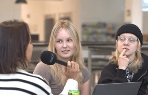 Drei junge Frauen sitzen in einem Haus. Eine Frau, die ein Mikrofon in der Hand hält, interviewt die beiden anderen. Eine der Befragten schaut die Sprecherin an und lächelt, während die andere, die eine Brille und eine Mütze trägt, ihre Lippen berührt und nachdenklich zuhört.