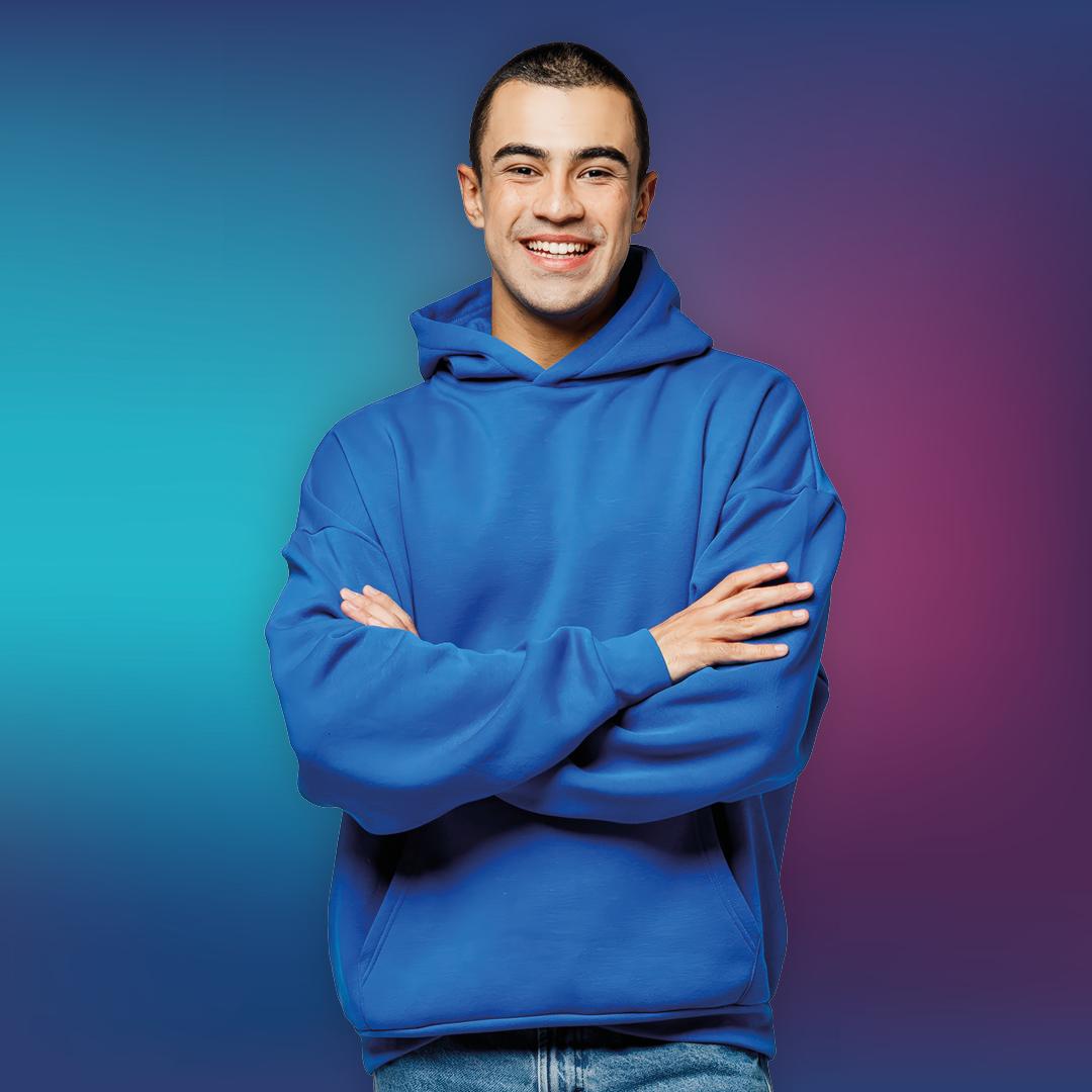 Eine junge Person mit kurzen dunklen Haaren, die einen blauen Kapuzenpulli und Jeans trägt, steht lächelnd mit verschränkten Armen vor einem blau-lila Hintergrund mit Farbverlauf.
