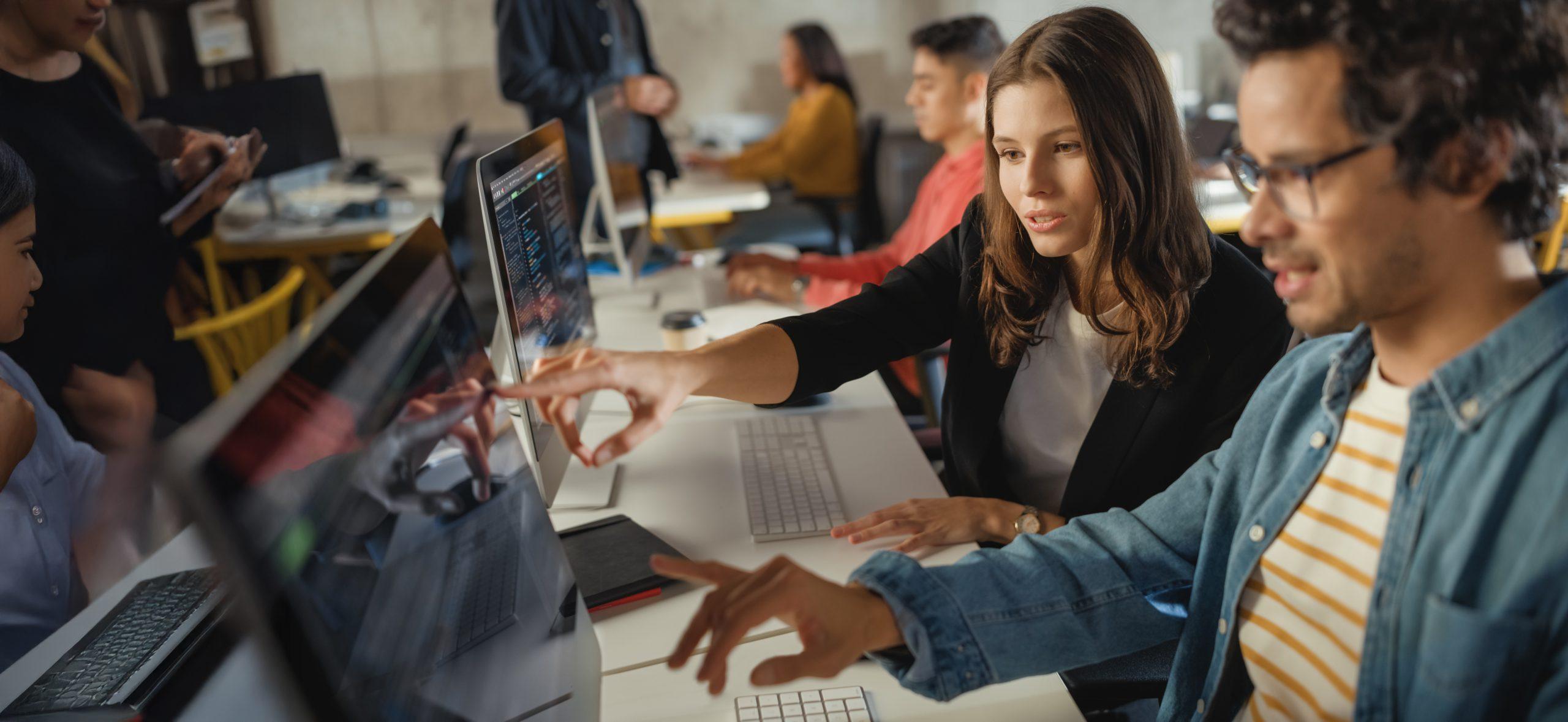 Eine Frau und ein Mann sitzen an einem Schreibtisch in einem Büro, konzentriert auf einen Computerbildschirm. Die Frau deutet auf den Bildschirm, während sie mit dem Mann etwas bespricht. Im Hintergrund arbeiten andere Kollegen an Computern.