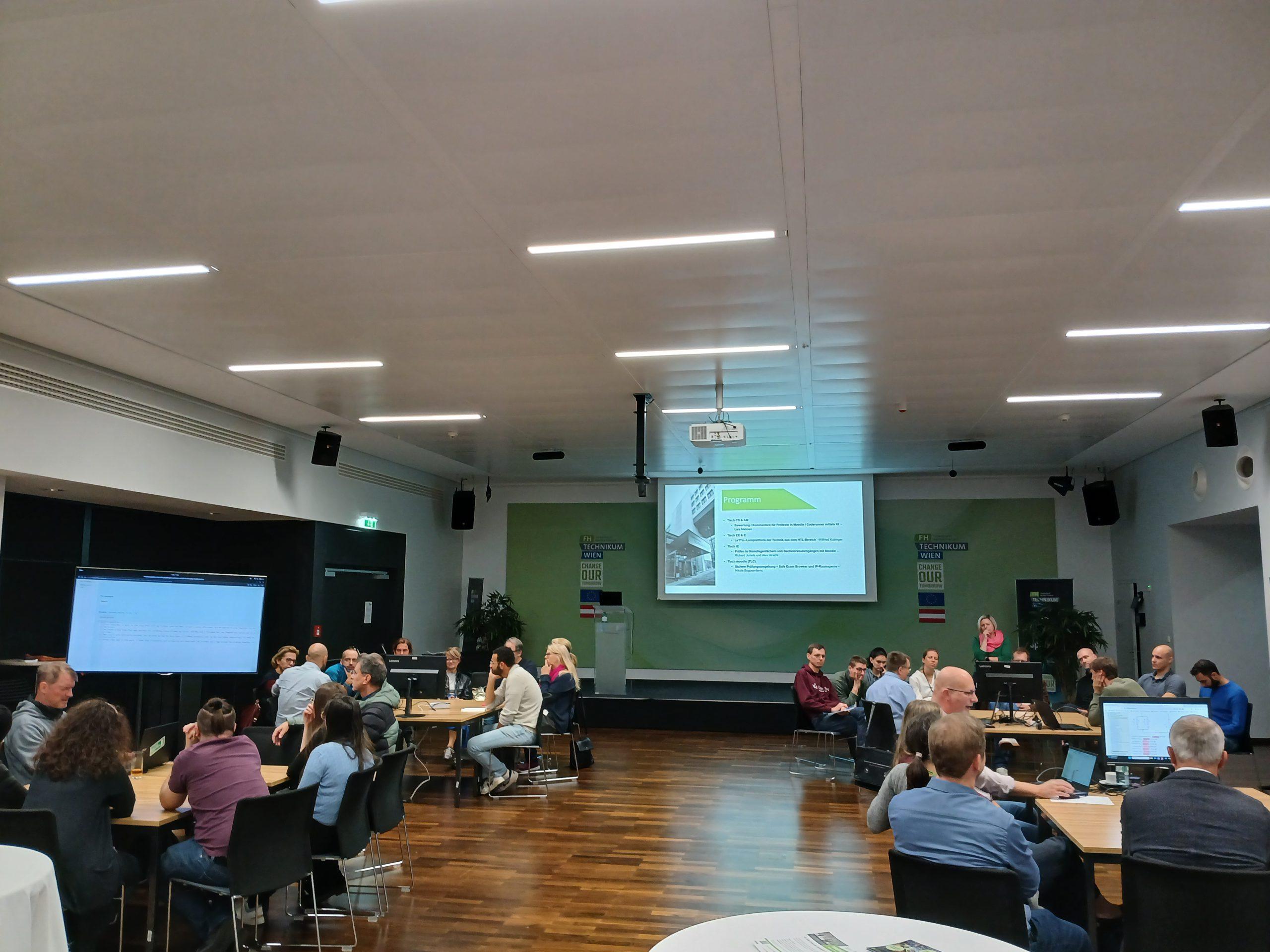 Eine Gruppe von Personen sitzt an runden Tischen in einem modernen Konferenzraum und sieht sich eine Präsentation an, die auf eine Leinwand im vorderen Teil des Raums projiziert wird. Einige sind in eine Diskussion vertieft, und auf einigen Tischen sind Laptops zu sehen.