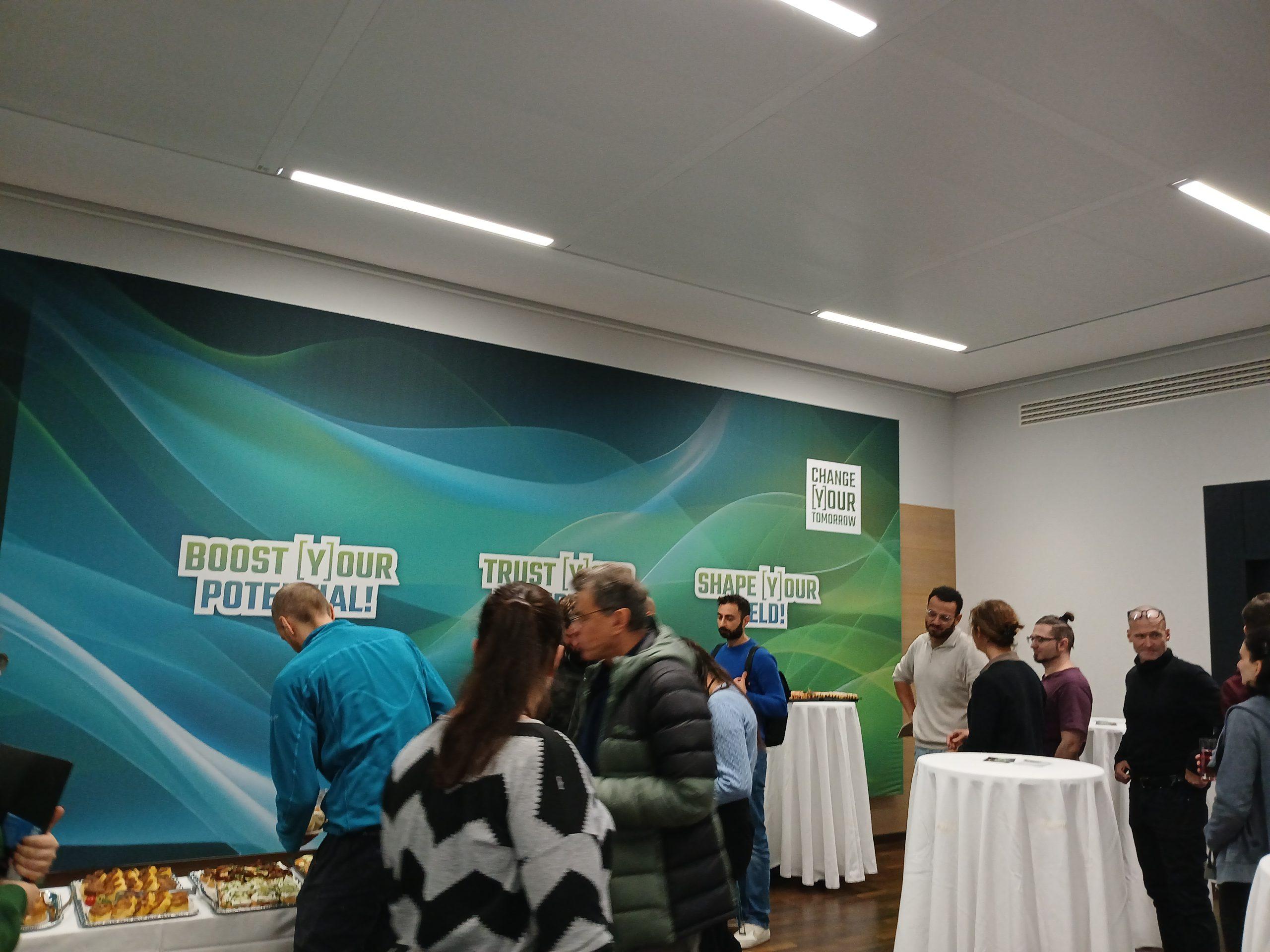 Eine Gruppe von Menschen versammelt sich um hohe Tische und ein Buffet in einem modernen Raum mit motivierenden Postern an der Wand, auf denen 'BOOST YOUR POTENTIAL', 'TRUST IN YOU' und 'SHAPE YOUR DESTINATION' steht.