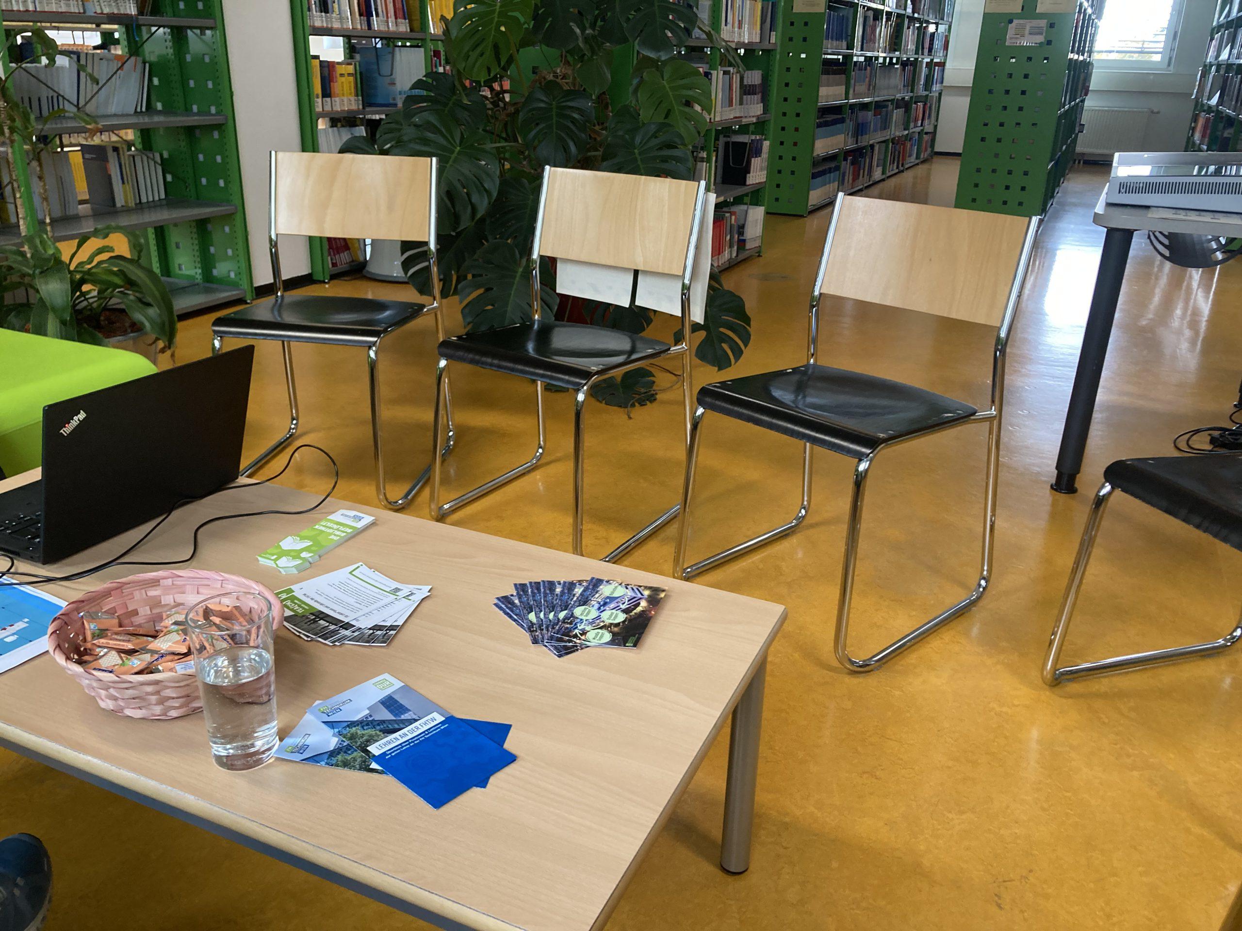 Eine Bibliotheksumgebung mit vier leeren Stühlen in einer Reihe, einem Holztisch mit Flyern, einem Glas Wasser, einem Korb mit Stiften und einem Laptop; im Hintergrund stehen Bücherregale und Grünpflanzen.