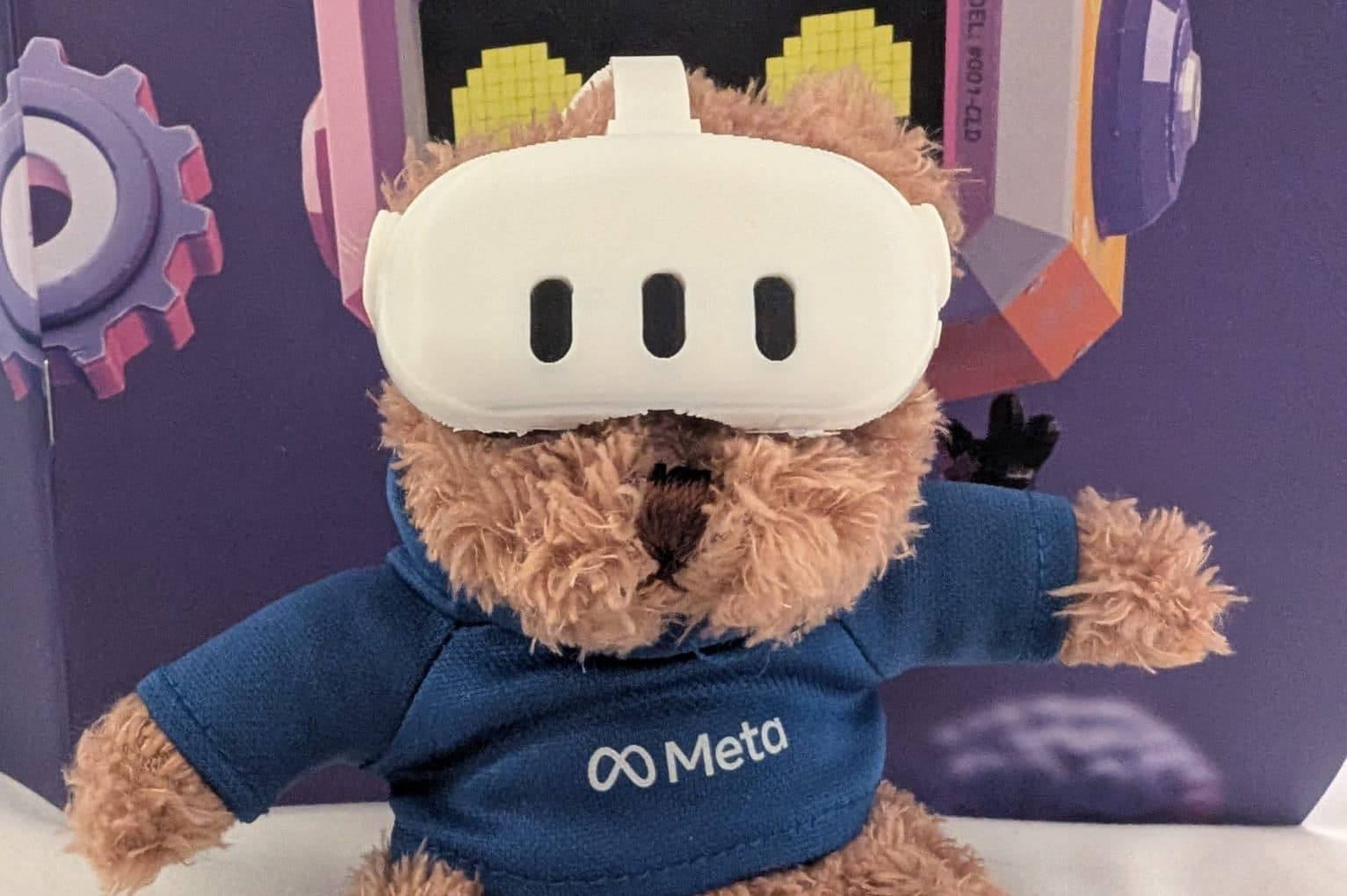 Ein brauner Teddybär, der einen blauen Kapuzenpullover mit dem Meta-Logo trägt, sitzt vor einem bunten Hintergrund. Der Bär hat ein weißes VR-Headset, das seine Augen bedeckt.