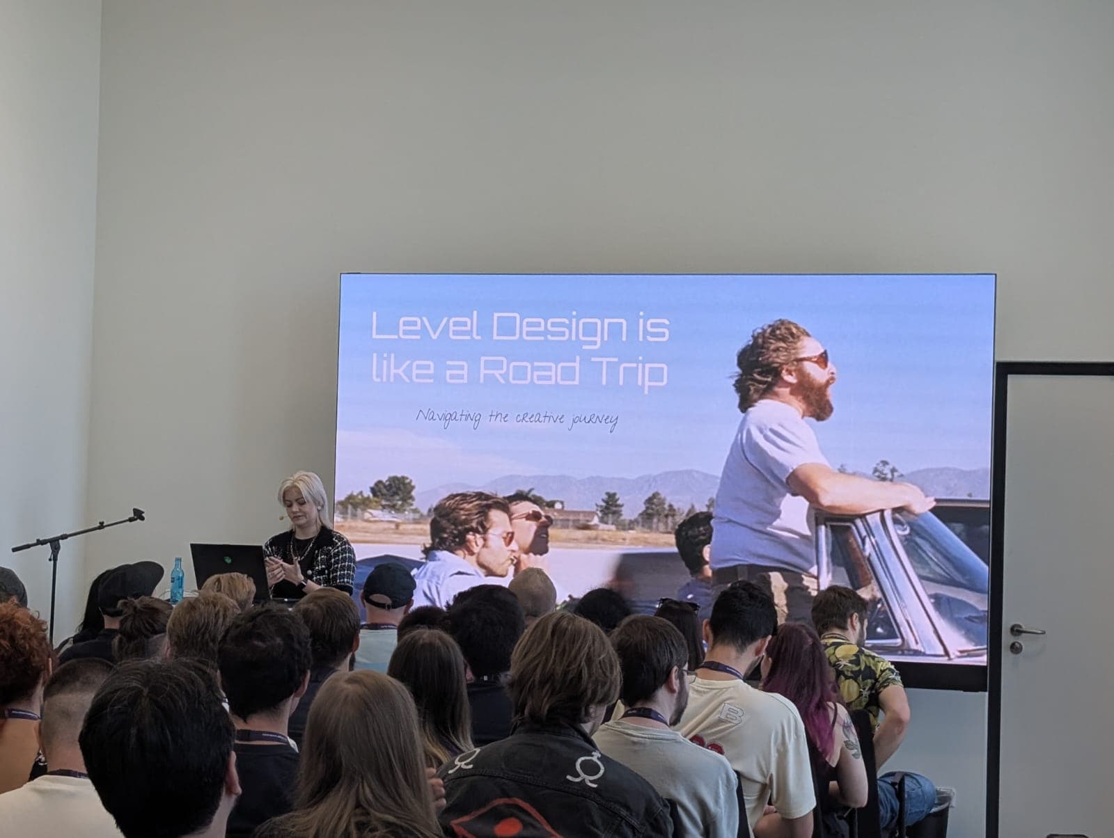 Eine Frau steht an einem Podium und spricht zu einem Publikum in einem Raum. Hinter ihr befindet sich ein großer Bildschirm, auf dem eine Präsentationsfolie mit dem Titel 'Level Design is like a Road Trip' zu sehen ist: Navigieren auf der kreativen Reise', mit einem Foto von Menschen in einem Cabrio.