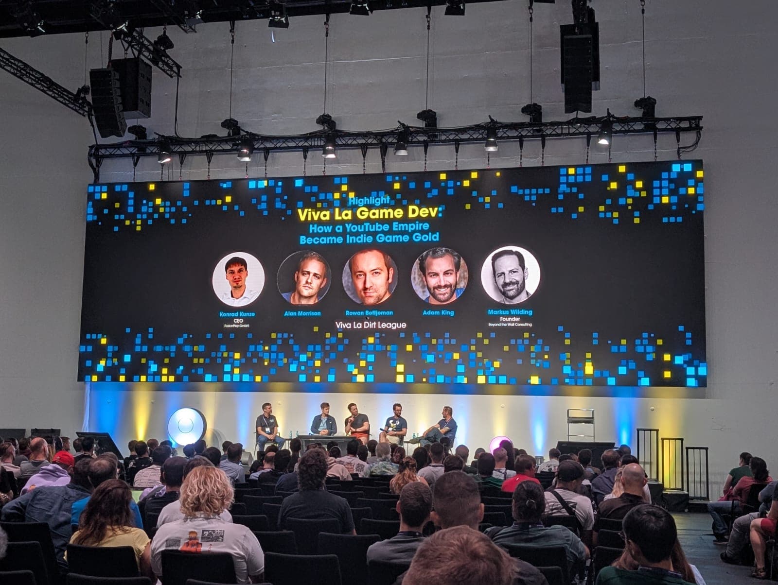 Eine Podiumsdiskussion mit dem Titel Viva La Game Dev findet auf der Bühne vor einem sitzenden Publikum statt. Auf einer großen Leinwand hinter den Rednern werden der Titel des Panels, das Thema sowie Fotos und Namen der vier Diskussionsteilnehmer angezeigt.