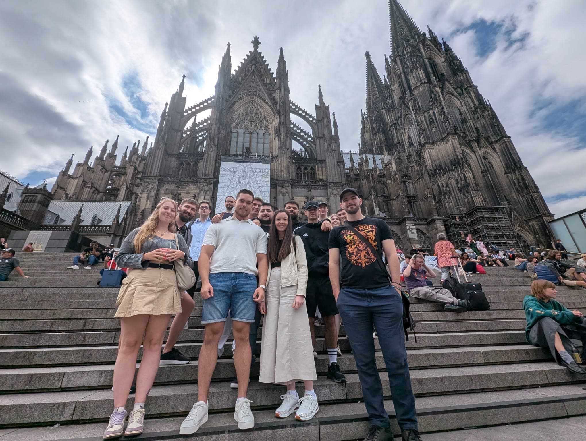 Eine Gruppe von Menschen posiert und lächelt auf den Stufen vor einer großen gotischen Kathedrale mit hohen Türmen und detaillierter Architektur; andere Besucher sitzen und gehen im Hintergrund unter einem bewölkten Himmel.