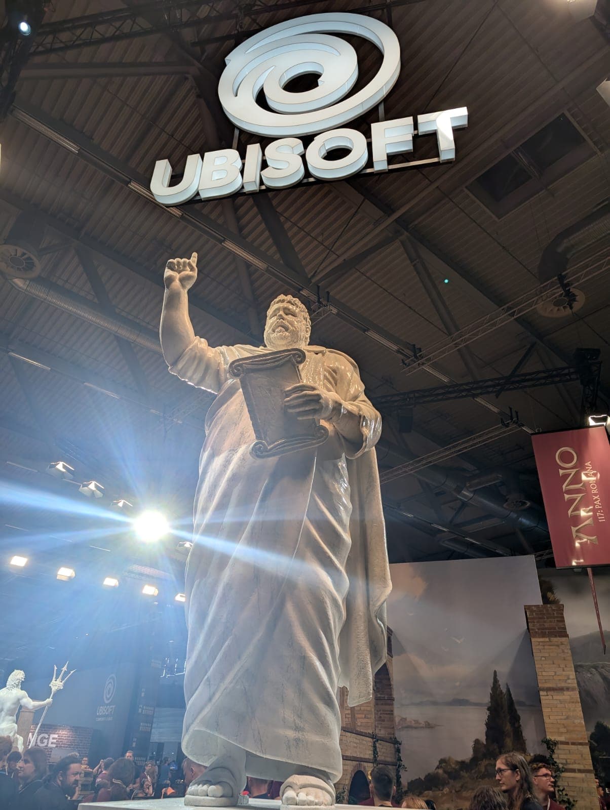 Große Statue einer gewandeten Figur, die eine Tafel hält und eine Hand hebt, ausgestellt unter einem auffälligen leuchtenden UBISOFT-Schild bei einer Indoor-Veranstaltung mit Scheinwerfern und Ausstellungsbannern.