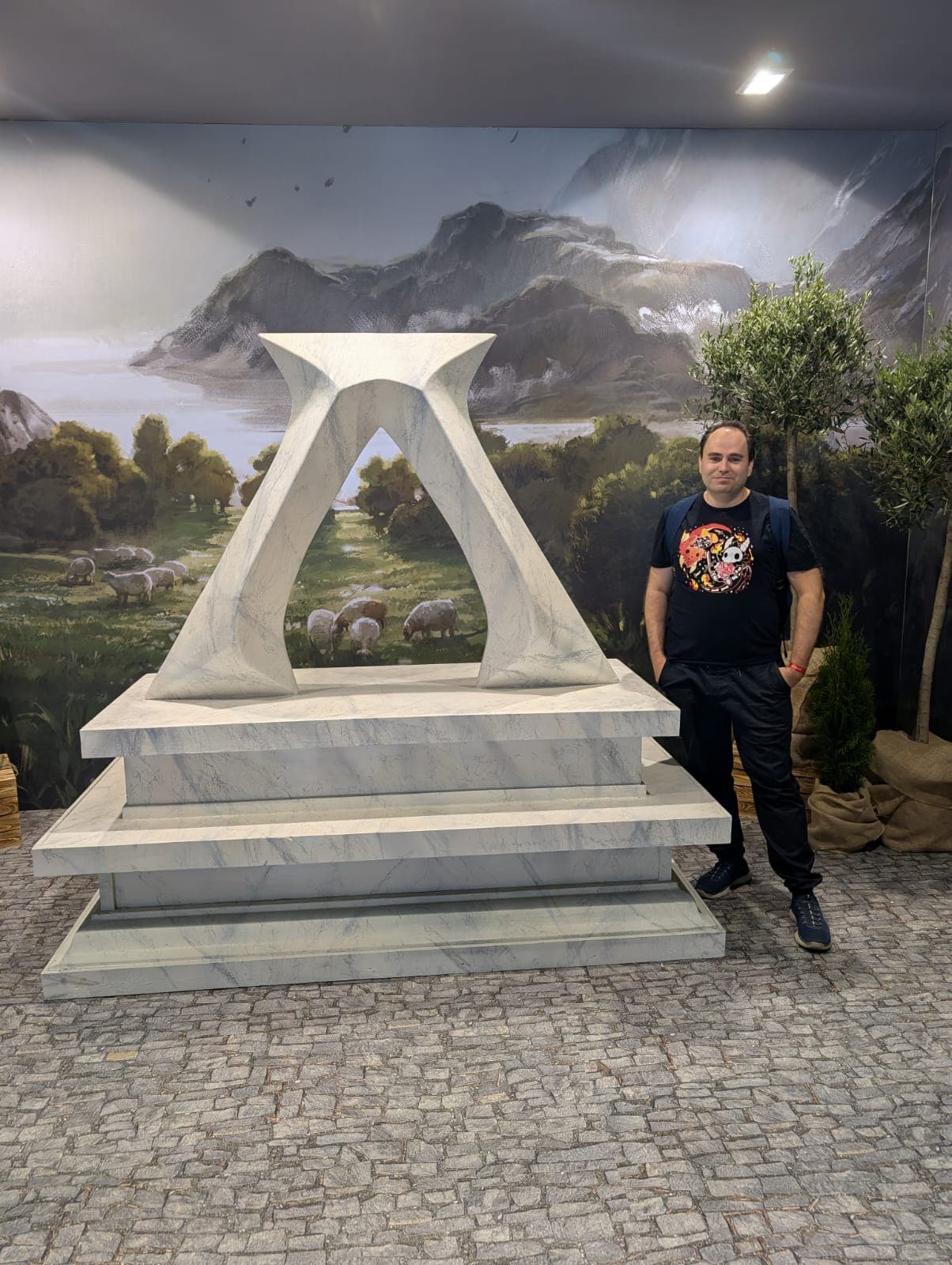 Ein Mann steht neben einer modernen weißen Marmorskulptur auf einem gestuften Sockel, mit einer Wandmalerei von Bergen, Bäumen und grasenden Schafen im Hintergrund. Der Boden besteht aus Kopfsteinpflaster, und in der Nähe stehen Topfpflanzen.