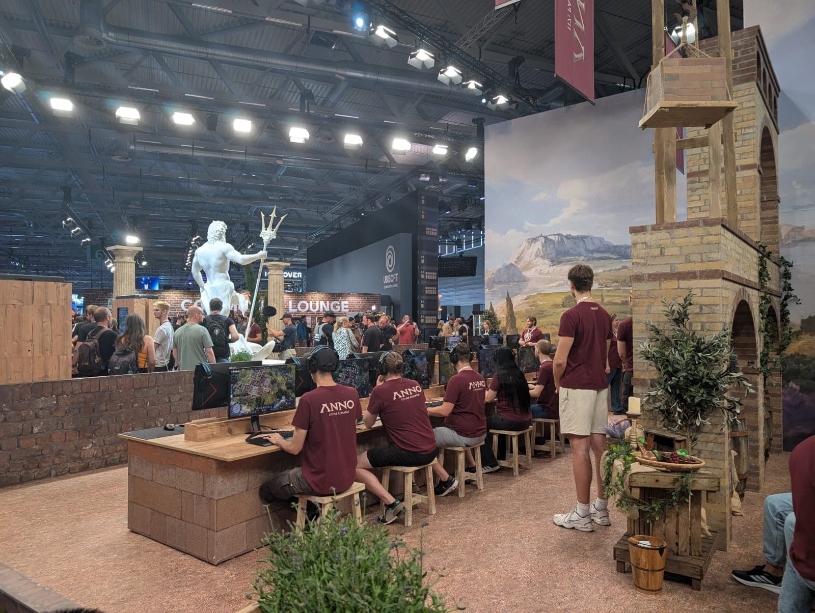Menschen in passenden kastanienbraunen Anno-Hemden spielen ein Videospiel an Holztischen in einem Veranstaltungsraum. Eine große Poseidon-Statue und ein Backsteinturm sind zu sehen, im Hintergrund eine Menschenmenge und Spruchbänder.