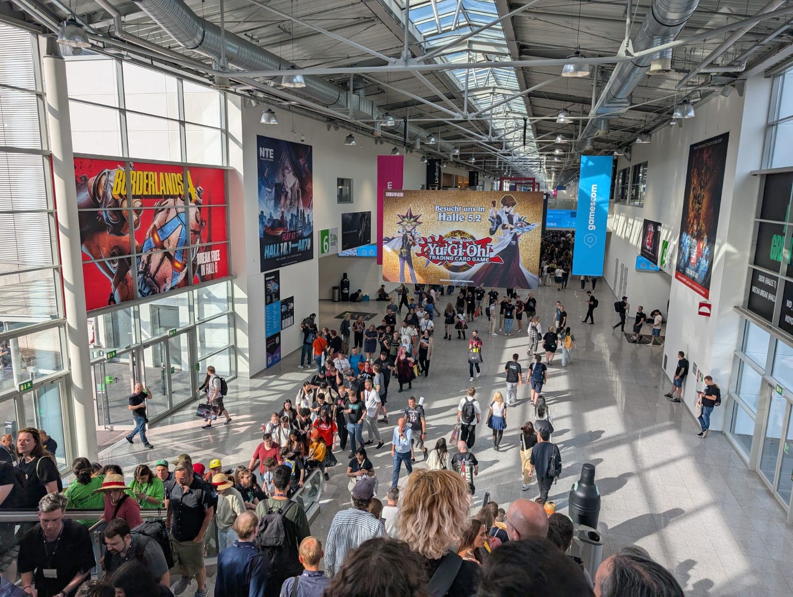 Eine große Menschenmenge läuft durch einen hell erleuchteten Flur des Convention Centers. An den Wänden hängen bunte Banner und Poster, darunter eines für Yu-Gi-Oh! Das Sonnenlicht fällt durch die hohen Fenster auf der linken Seite ein.