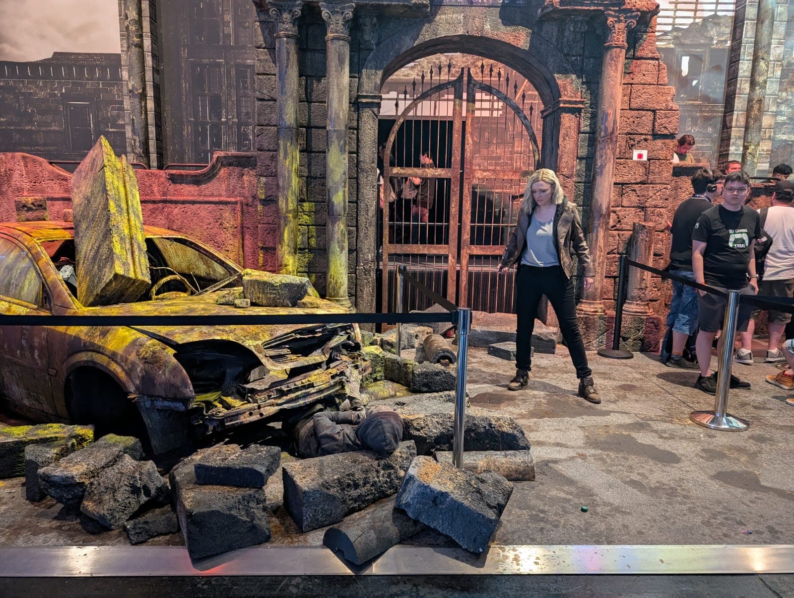 Eine Frau in einer schwarzen Jacke steht neben einem beschädigten, moosbewachsenen Auto und zerbröckelten Ziegelsteinen in einer thematischen Ausstellung, während Menschen hinter einer Absperrung zusehen. Im Hintergrund ist eine Steinstruktur mit Tor zu sehen.