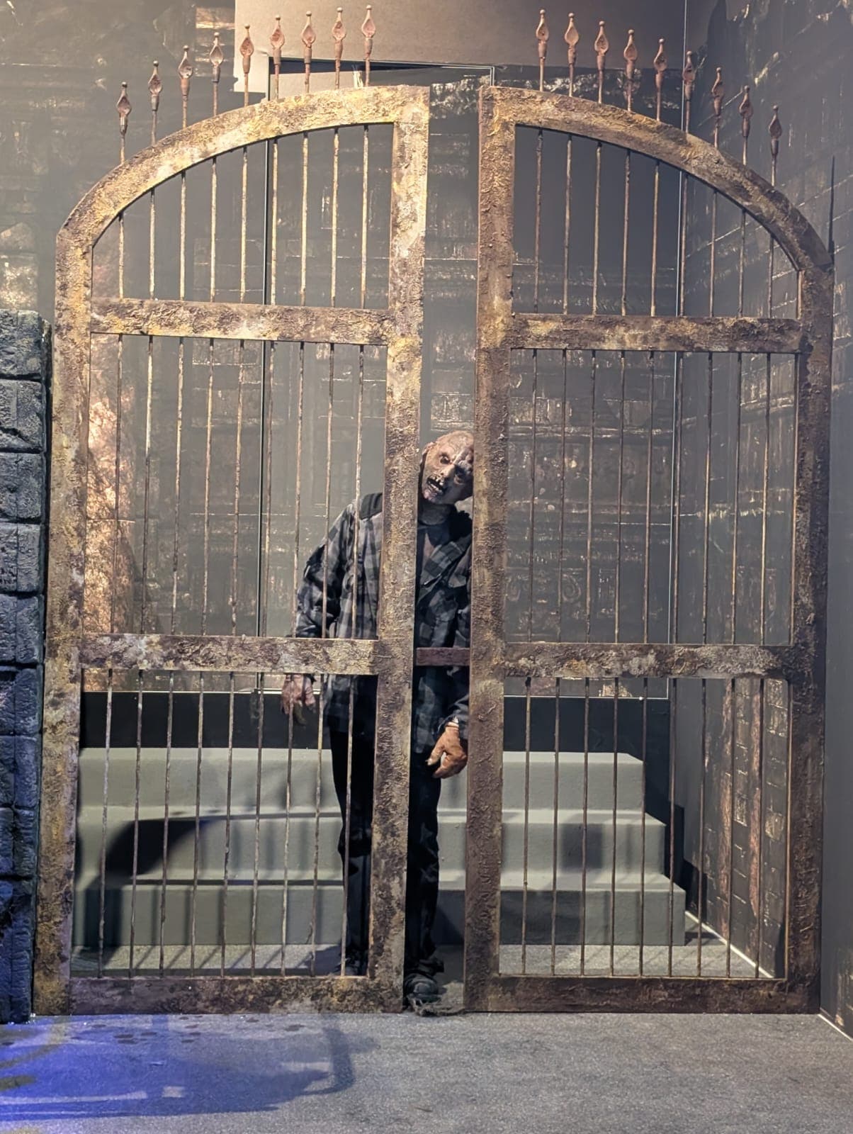Eine als Zombie verkleidete Person steht hinter einem großen, verwitterten Eisentor mit spitzen Spitzen und lehnt sich zur Seite. Der Hintergrund besteht aus Steinmauern und Stufen, die eine dunkle, unheimliche Atmosphäre schaffen.