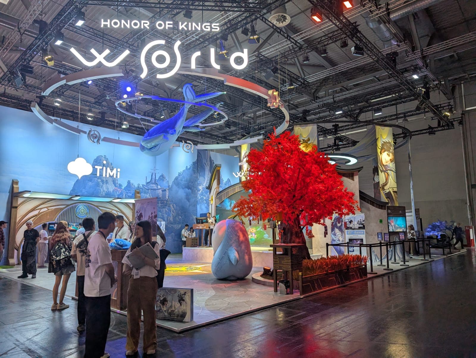 Messestand für Honor of Kings: World at an event, mit einem großen roten künstlichen Baum, einem blauen fliegenden Kreaturmodell darüber, Fantasy-Dekoration und Menschen, die im Ausstellungsbereich interagieren.