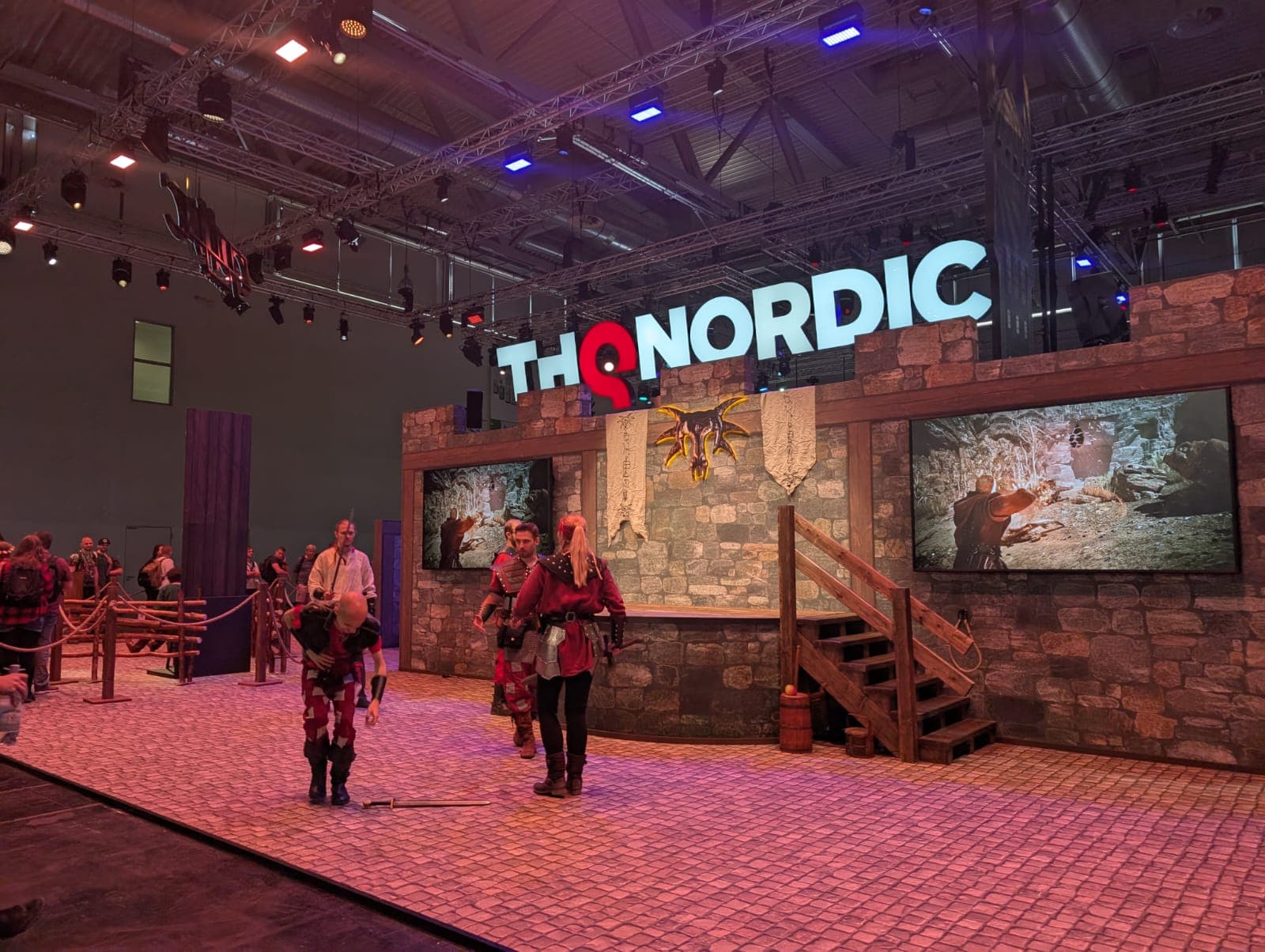 Ein Messestand von THQ Nordic zeigt eine mittelalterliche Kulisse mit Steinwänden, Holztreppen, Bannern und zwei großen Bildschirmen. Cosplayer in Fantasiekostümen interagieren mit den Besuchern auf der Ausstellungsfläche.