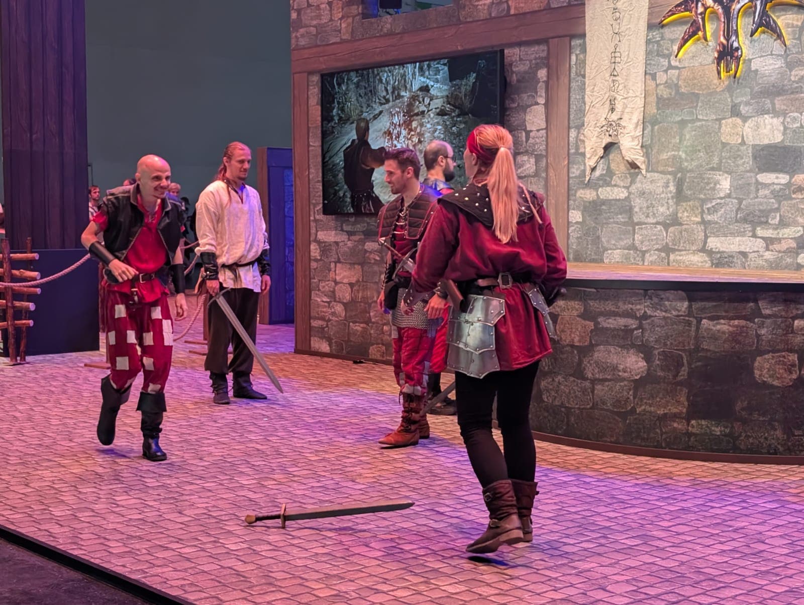 Fünf Personen in mittelalterlichen Kostümen proben einen Schwertkampf auf einer mit Steinen gepflasterten Bühne vor einer Burgkulisse. Ein Schwert liegt auf dem Boden, und ein Monitor zeigt eine Fantasieszene an der Wand hinter ihnen.