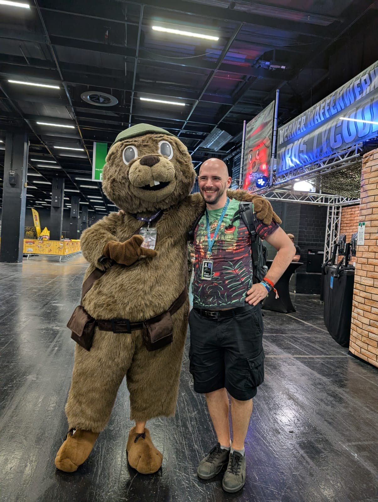 Eine Person in einem großen Otterkostüm steht mit dem Arm um einen lächelnden Mann, der ein bedrucktes T-Shirt und Shorts trägt, auf einer Indoor-Konferenz oder -Veranstaltung, mit Ständen und Schildern im Hintergrund.