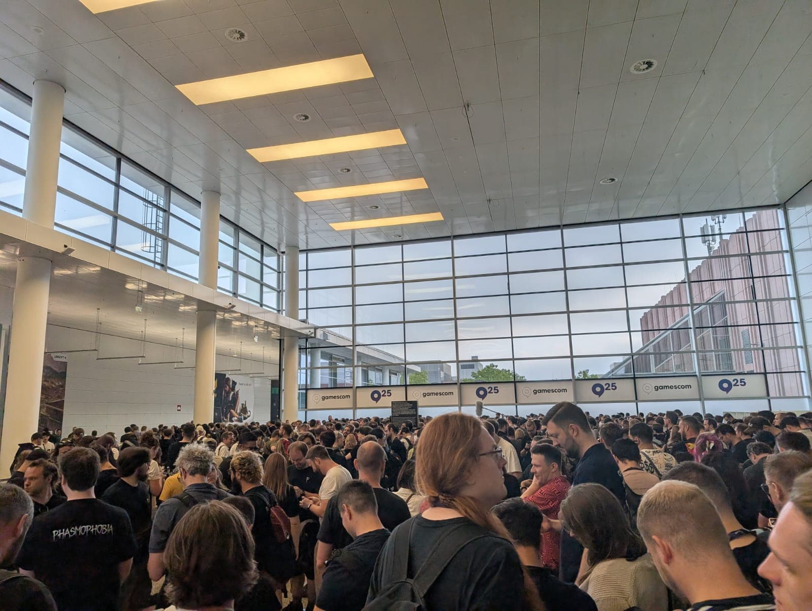 Eine große Menschenmenge füllt eine geräumige, moderne Halle mit hohen Decken und großen Fenstern. Darüber hängen Schilder mit den Aufschriften 'gamescom' und 'Q25'. Die Atmosphäre ist geschäftig und lebhaft.