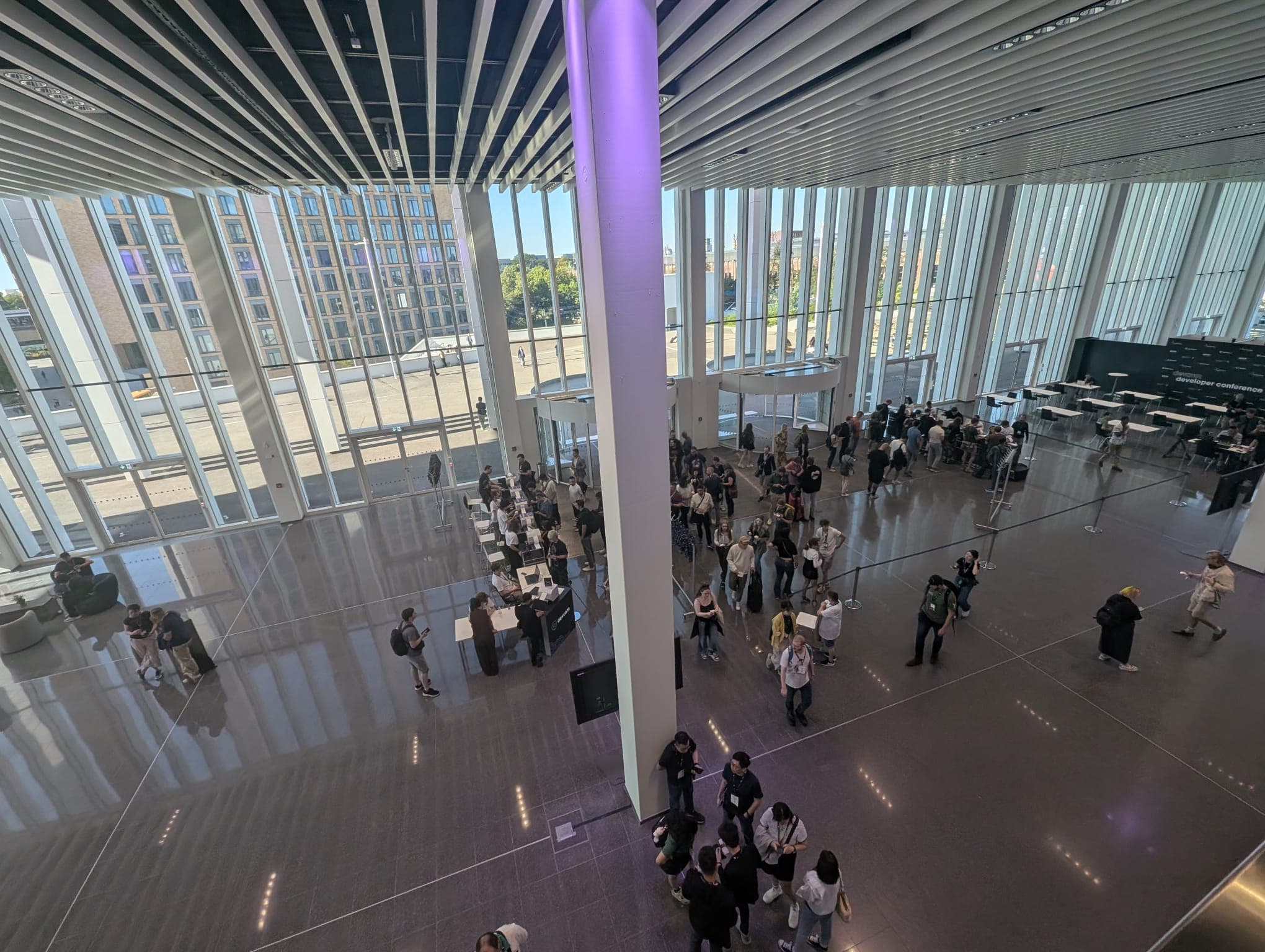 Ein geräumiges Atrium mit hohen Fenstern, durch die natürliches Licht einfällt, auf dessen glänzendem Boden sich Gruppen von Menschen versammeln. An der Seite sind einige Tische und Stühle aufgestellt, und von draußen kann man eine Stadtlandschaft sehen.