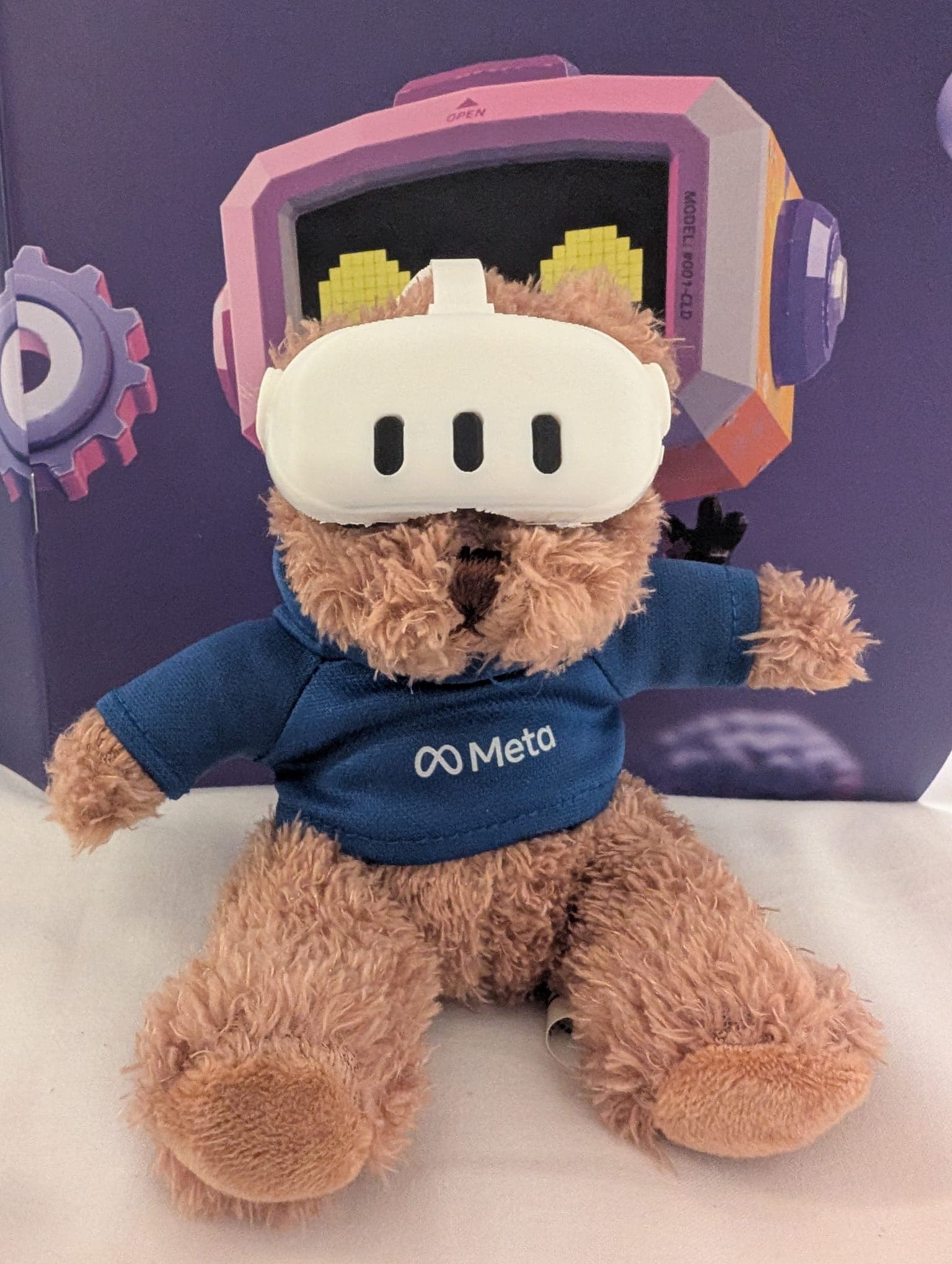 Ein brauner Teddybär, der einen blauen Meta-Kapuzenpulli und ein weißes VR-Headset trägt, sitzt vor einem violetten Hintergrund, auf dem ein Cartoon-Roboter mit verpixelten gelben Augen zu sehen ist.