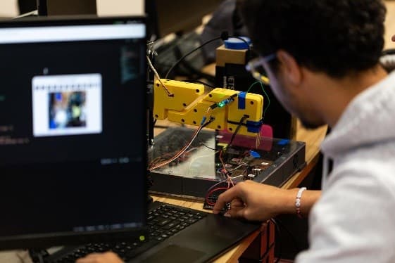 Eine Person arbeitet an einem Laptop neben einem gelben Roboterarm und interagiert mit angeschlossenen Drähten und elektronischen Komponenten auf einem Schreibtisch in einer Laborumgebung.