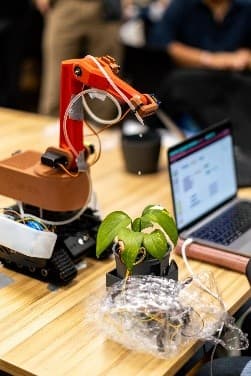 Ein Roboterarm gießt eine kleine Topfpflanze auf einem Holztisch neben einem Laptop, im Hintergrund sind unscharfe Menschen zu sehen.