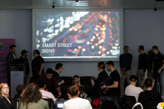 Eine Gruppe von Menschen ist in einem Raum versammelt, einige stehen, andere sitzen, vor einem großen Bildschirm, auf dem eine verschwommene Stadtlandschaft und die Worte SMART STREET SIGNS zu sehen sind.