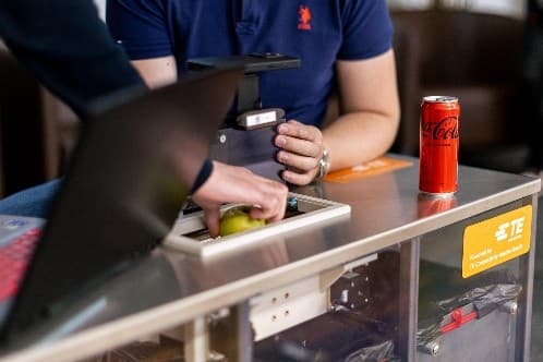 Eine Person legt einen grünen Apfel in ein kleines scannerähnliches Gerät, während eine andere den Deckel festhält. Eine rote Coca-Cola-Dose liegt auf dem Tisch daneben. Der Aufbau scheint Teil einer Technologievorführung oder eines Experiments zu sein.
