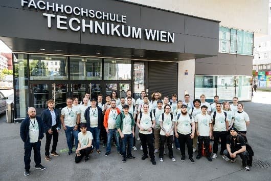 Eine Gruppe von Menschen in hellen Hemden steht an einem sonnigen Tag vor dem Gebäude der Fachhochschule Technikum Wien und stellt sich für ein Gruppenfoto auf.