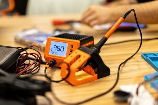 Eine digitale Lötstation mit einer orangefarbenen Anzeige, die 360 °C anzeigt, ein Lötkolben in seiner Halterung und Drähte auf einem Holztisch. Die Hände einer Person sind im Hintergrund verschwommen.