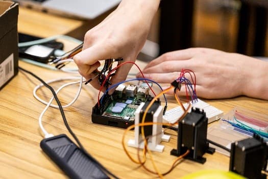 Die Hände einer Person arbeiten mit Drähten und elektronischen Bauteilen, die mit einer Leiterplatte auf einem Holztisch verbunden sind, und bauen ein elektronisches Projekt zusammen oder suchen nach Fehlern.