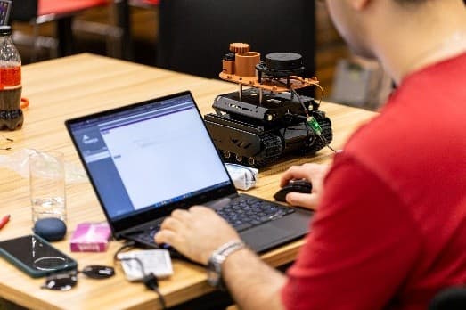 Eine Person, die einen Laptop neben einem kleinen Roboterfahrzeug auf einem Holztisch benutzt, mit diversem elektronischen Zubehör und einer Getränkeflasche in der Nähe.