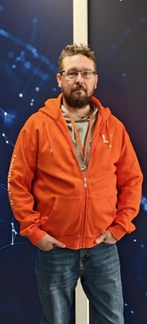 Ein Mann mit kurzen Haaren, Brille und Bart trägt einen orangefarbenen Kapuzenpulli mit Reißverschluss über einem grauen Hemd und blauen Jeans. Er steht vor einem blauen Hintergrund mit weißen abstrakten Linien und Punkten, die Hände in den Taschen.