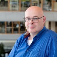 Ein Mann mittleren Alters mit Brille und Glatze, der ein blaues Hemd trägt, steht in einem Innenraum vor einem unscharfen Bürohintergrund.