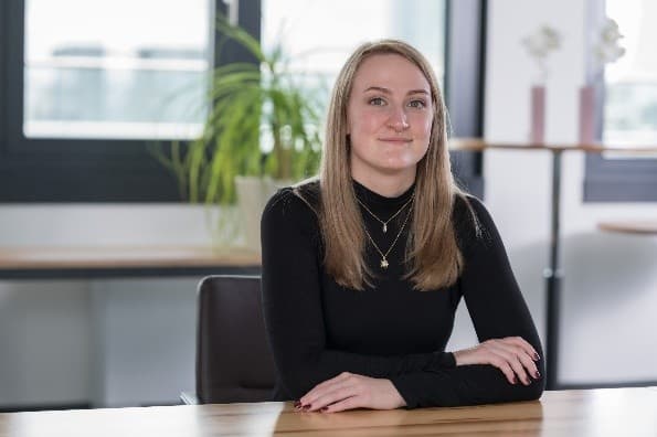 Eine Frau mit glattem blondem Haar, die ein schwarzes langärmeliges Oberteil und mehrlagige Ketten trägt, sitzt mit gefalteten Händen an einem Schreibtisch. Im Hintergrund sind eine Pflanze und große Fenster zu sehen.