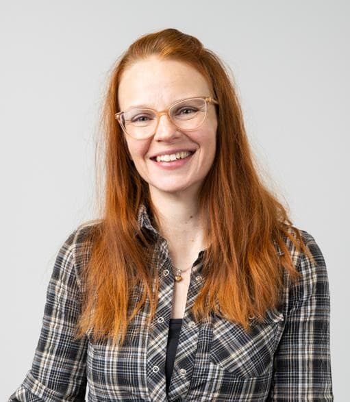 Eine Frau mit langen roten Haaren und Brille lächelt, trägt ein kariertes Hemd und eine Halskette und steht vor einem schlichten hellgrauen Hintergrund.