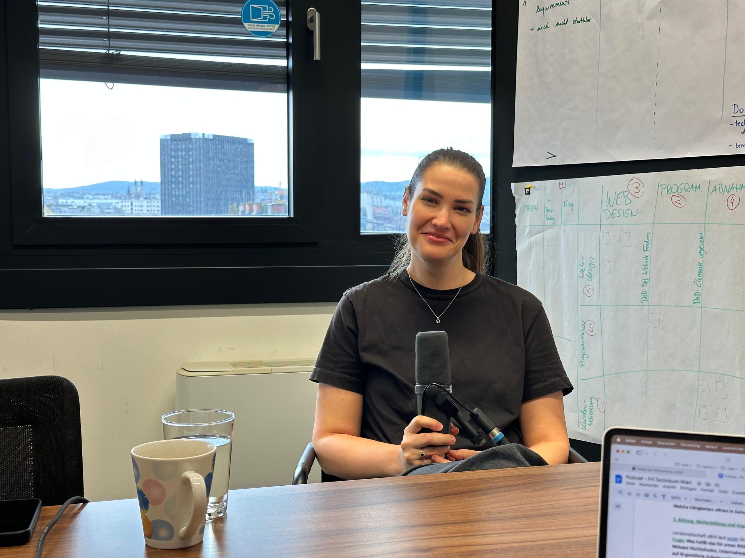 Eine Frau sitzt an einem Tisch in einem Büro, hält ein Mikrofon in der Hand und lächelt. Eine Kaffeetasse, ein Glas Wasser und ein Laptop stehen auf dem Tisch. Im Hintergrund sind große Fenster und Tabellen an der Wand zu sehen.