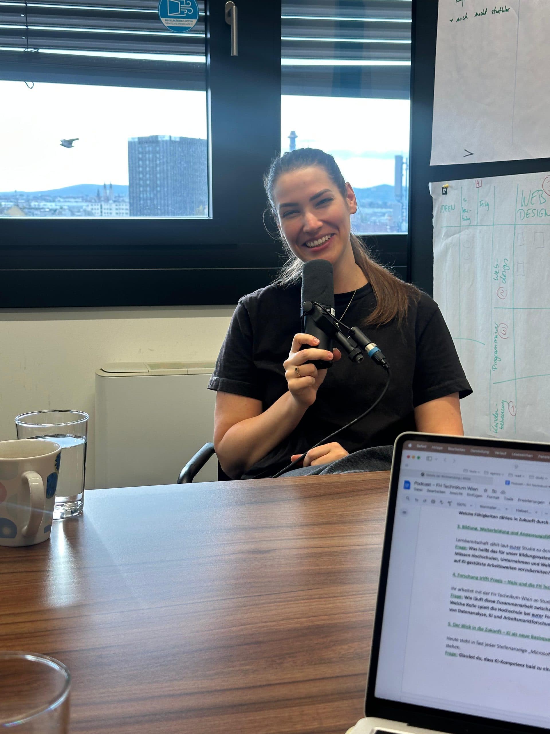 Eine Frau lächelt, während sie ein Mikrofon in einer Büroumgebung hält. Sie sitzt an einem Tisch mit einem Glas Wasser, einer Tasse und einem aufgeklappten Laptop. Hinter ihr befindet sich ein Fenster mit einer Stadtlandschaft und an der Wand befestigten Papieren.