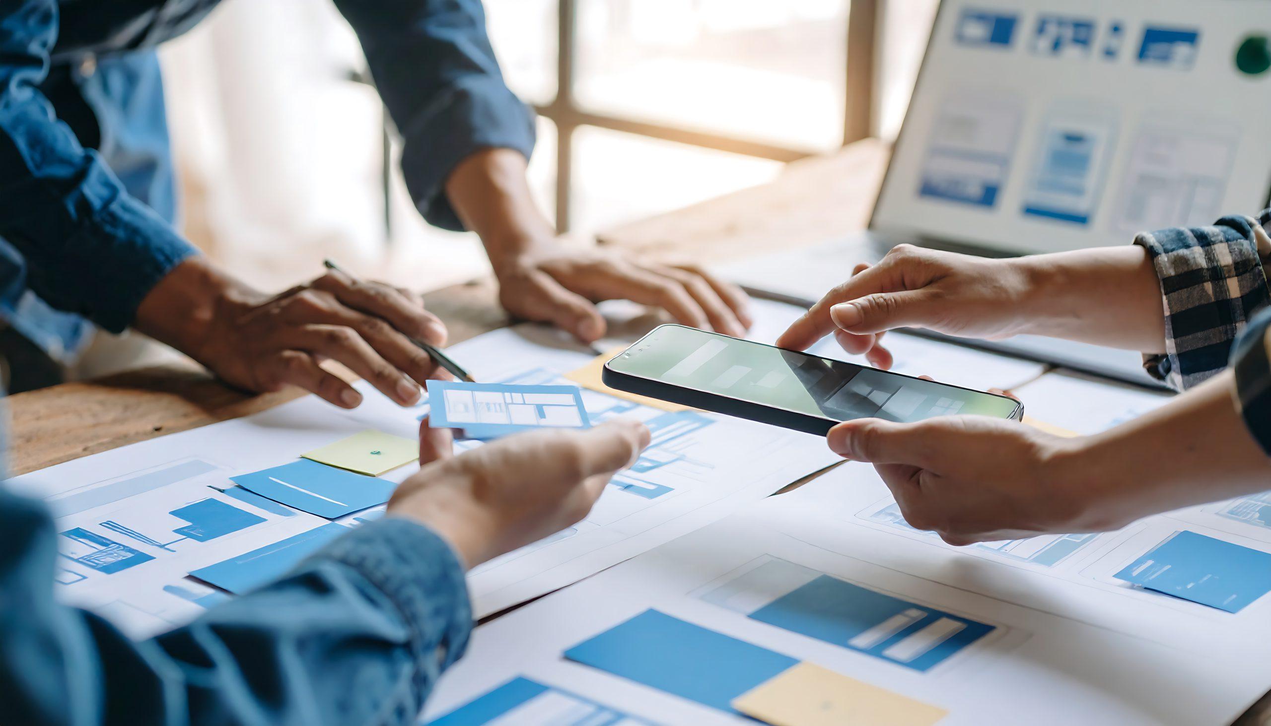 Drei Personen arbeiten an einem Schreibtisch zusammen, um gedruckte Web- oder App-Entwürfe zu prüfen und zu diskutieren. Eine Person hält ein Smartphone in der Hand, eine andere zeigt auf ein Papier, während ein Laptop im Hintergrund einen ähnlichen Entwurf anzeigt.